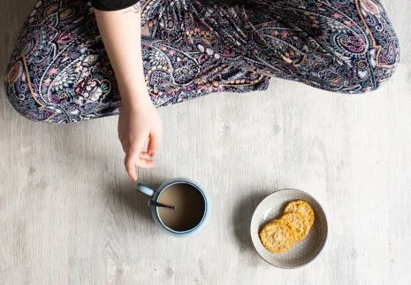 Person sitzt mit Kaffee und Keksen im Schneidersitz auf hellgrauem Fußboden in Holzoptik