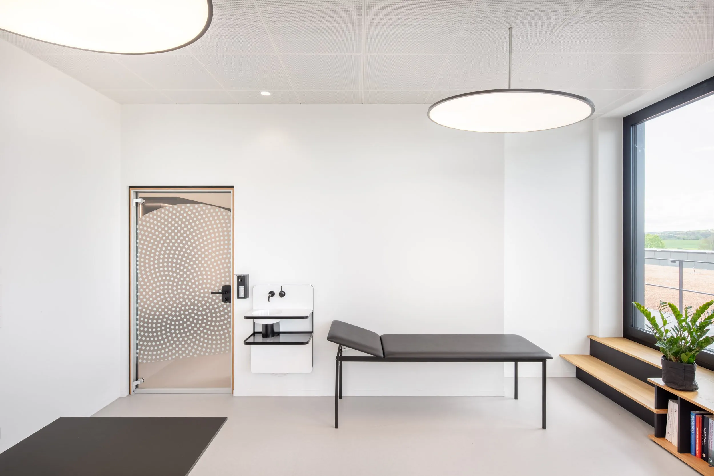 Modernes Behandlungszimmer mit großen Fenstern und einer medizinischen Liege auf grauem Betonboden
